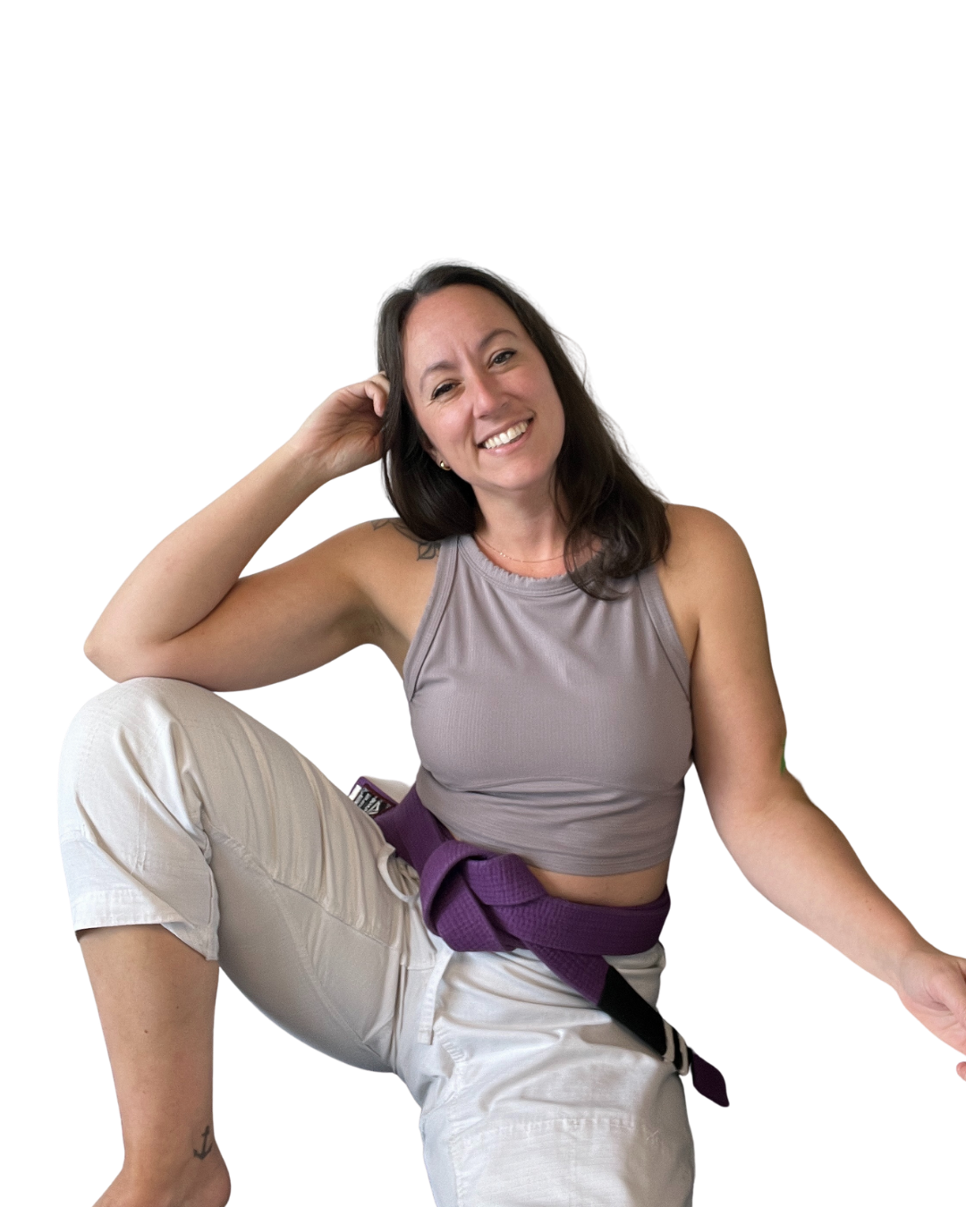 A woman with long dark hair sitting on the floor, smiling, wearing a gray crop top and white pants with a purple belt.