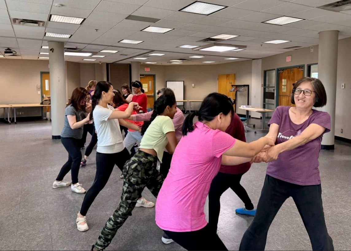 A group of women engaging in a game of tug-of-war indoors, with some smiling and others focused, in a spacious, well-lit room with light-colored walls and ceiling tiles.