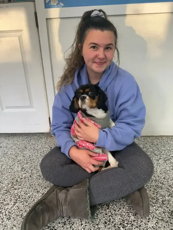 A young woman with curly hair tied with a bow, sitting cross-legged on the floor and holding a small tricolor puppy inside a comfortable dog boarding space in Omaha.