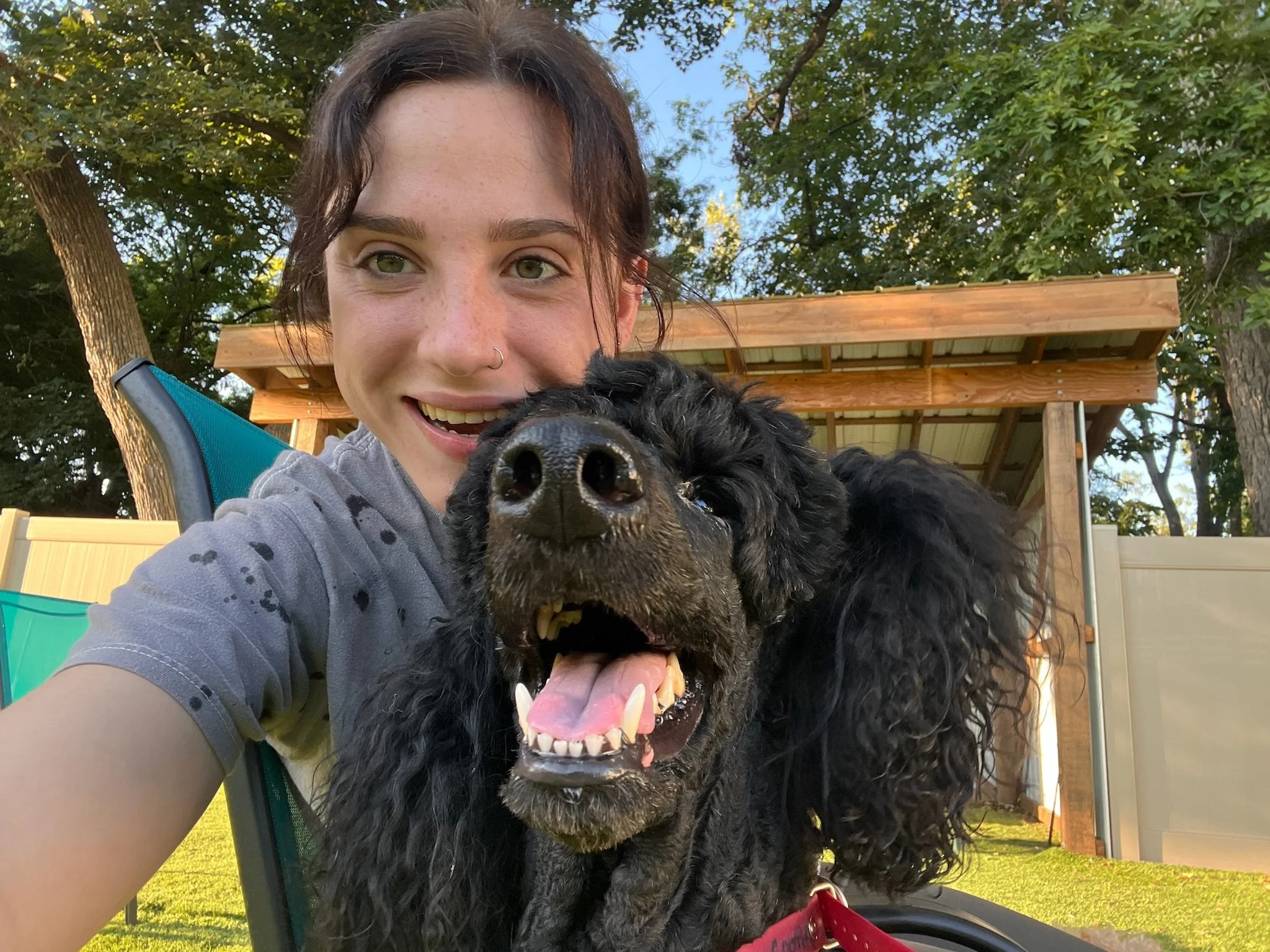 A woman and a black dog taking a selfie outdoors on a sunny day with trees and a wooden shed at Katie’s Kennel in Omaha, NE