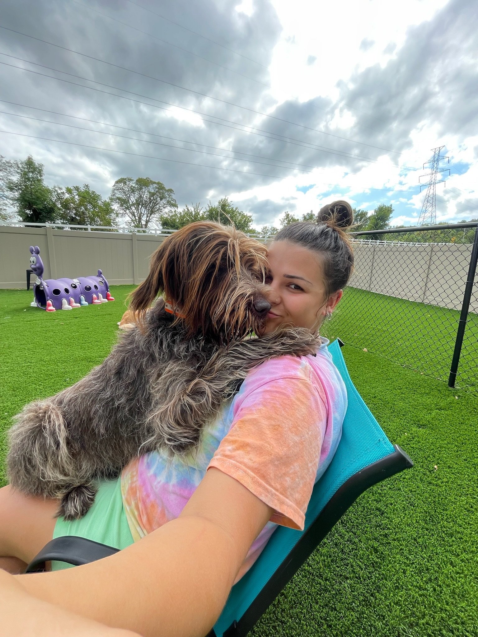 Young woman with dark hair holding a long-haired dog while sitting on a blue chair in a fenced backyard at Katie’s Kennel in Omaha, NE