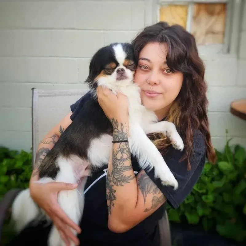 A woman with long wavy hair and tattoos hugging a small tricolor dog outside near a brick wall and plants at an Omaha dog boarding center.