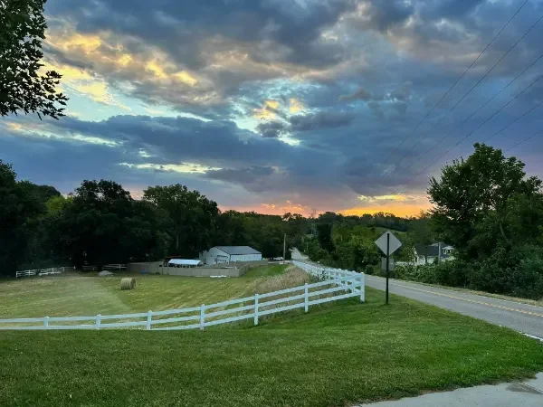 Sunset over a rural neighborhood with a white fence along a grassy yard, trees, a small building with a metal roof, and power lines at Katie's Kennel dog boarding in Omaha, NE.