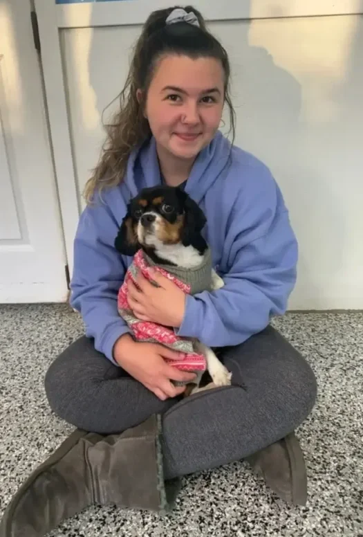 A young woman with a high ponytail wearing a light blue hoodie and gray leggings sitting cross-legged on a speckled floor, holding a small black, white, and tan puppy in her arms, both looking at the camera at Katie’s Kennel in Omaha, NE