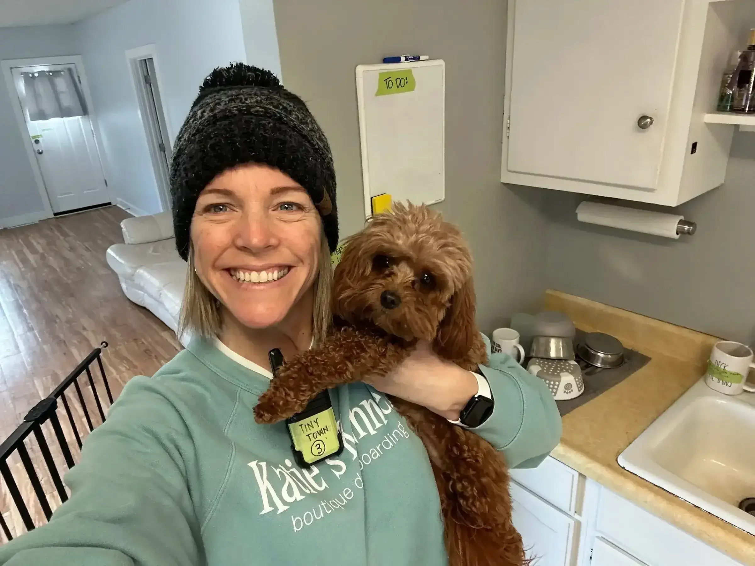 A smiling woman in a kitchen holding a curly-haired brown dog during a home-style moment connected to dog boarding in Omaha.
