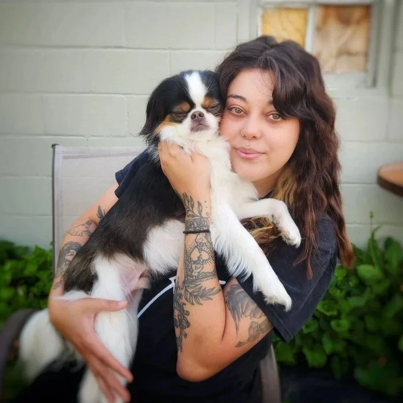 A woman with long wavy hair and tattoos hugging a small tricolor dog outside near a brick wall and plants at an Omaha dog boarding center.