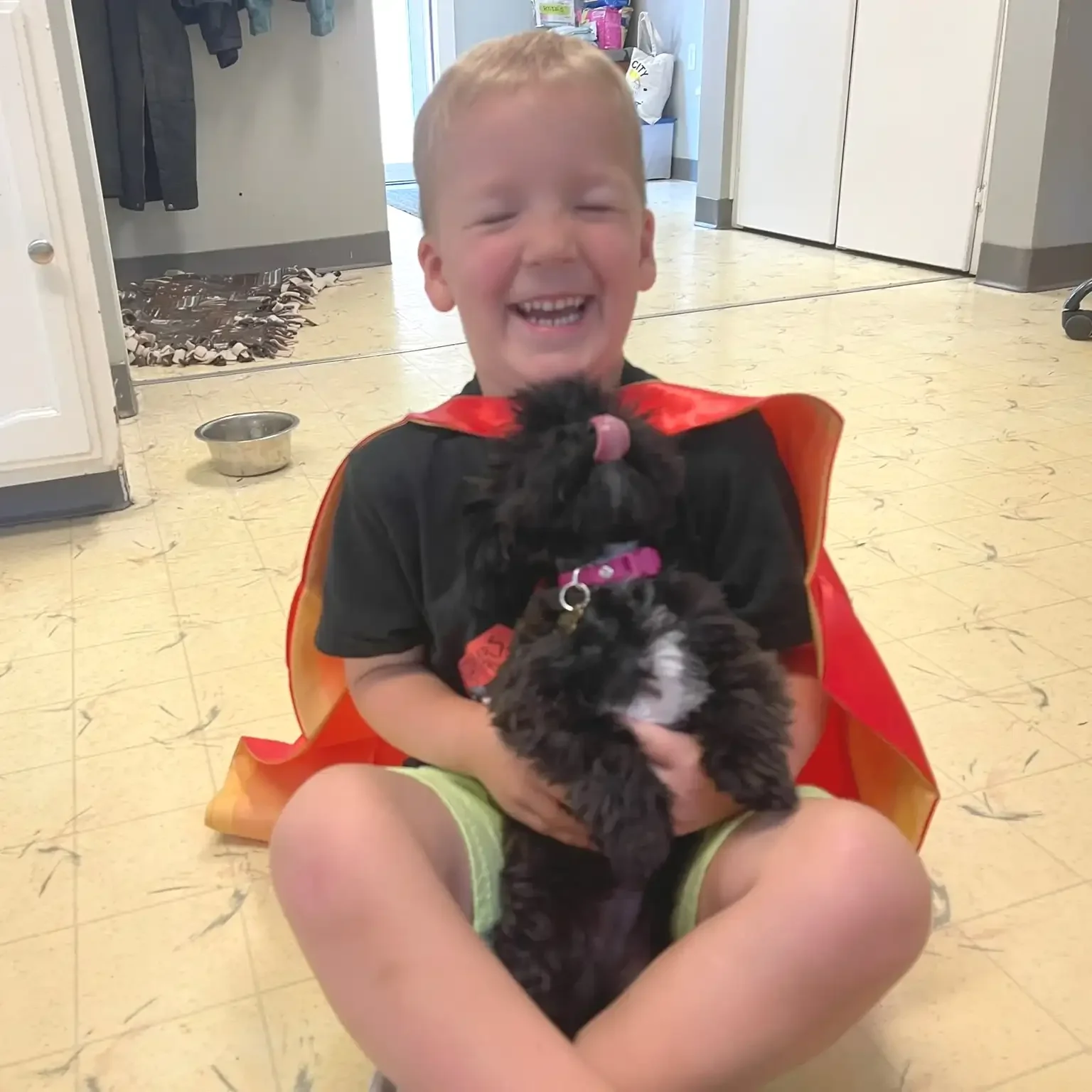 A young boy laughing while holding a black and white puppy indoors during a visit to a dog boarding facility.