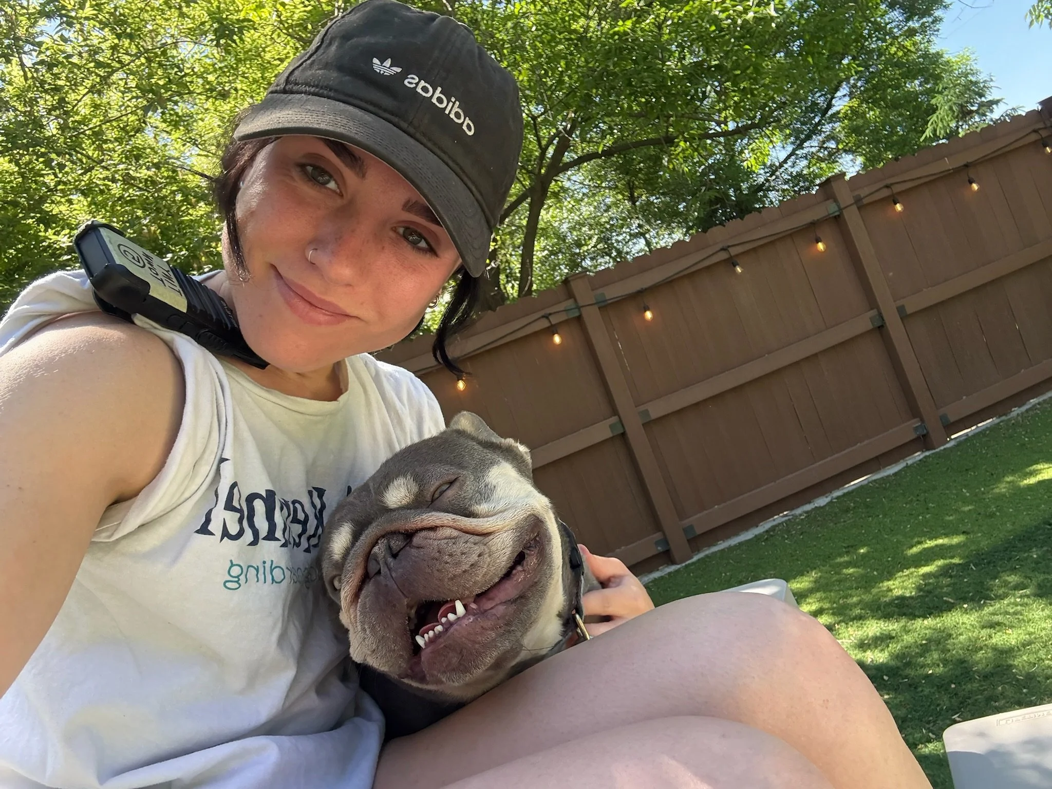 A woman smiling while taking a selfie with her happy bulldog in a backyard with a wooden fence and string lights hanging above, under a tree at a dog boarding facility in Omaha, NE