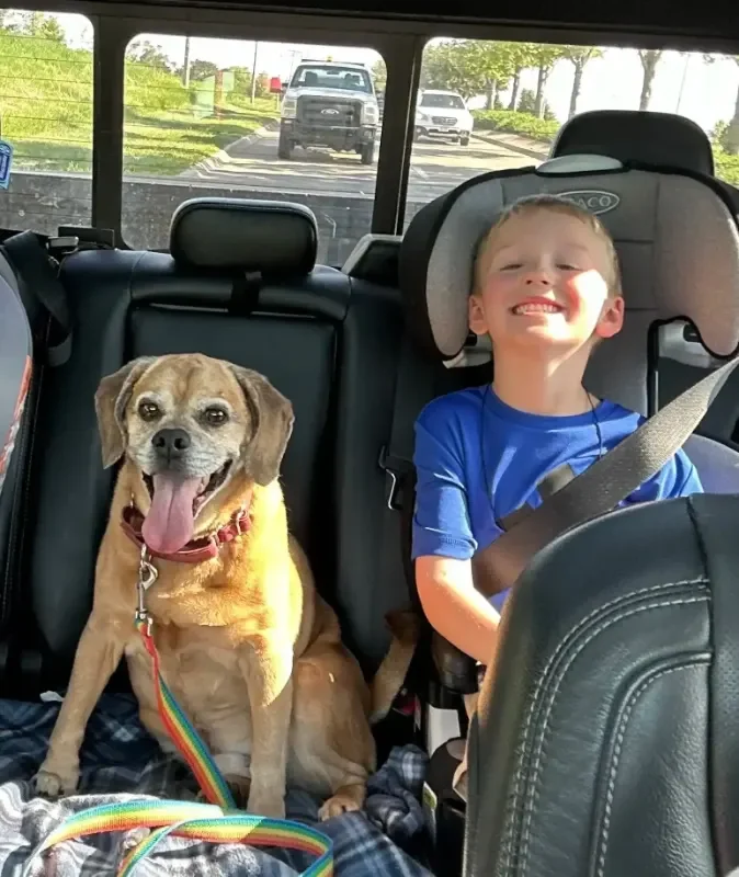 A smiling young boy sitting in a car seat with a happy dog beside him after pickup from an Omaha dog boarding stay.