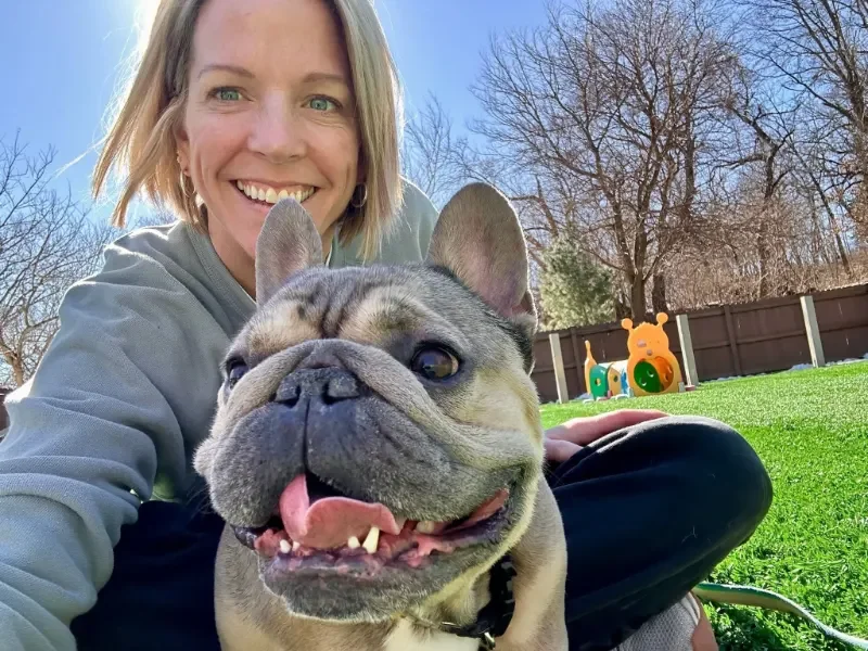A woman with short blonde hair smiling next to a happy bulldog with its tongue out, outdoors on a sunny day with trees and a fence in the background of a dog boarding facility in Omaha, NE.
