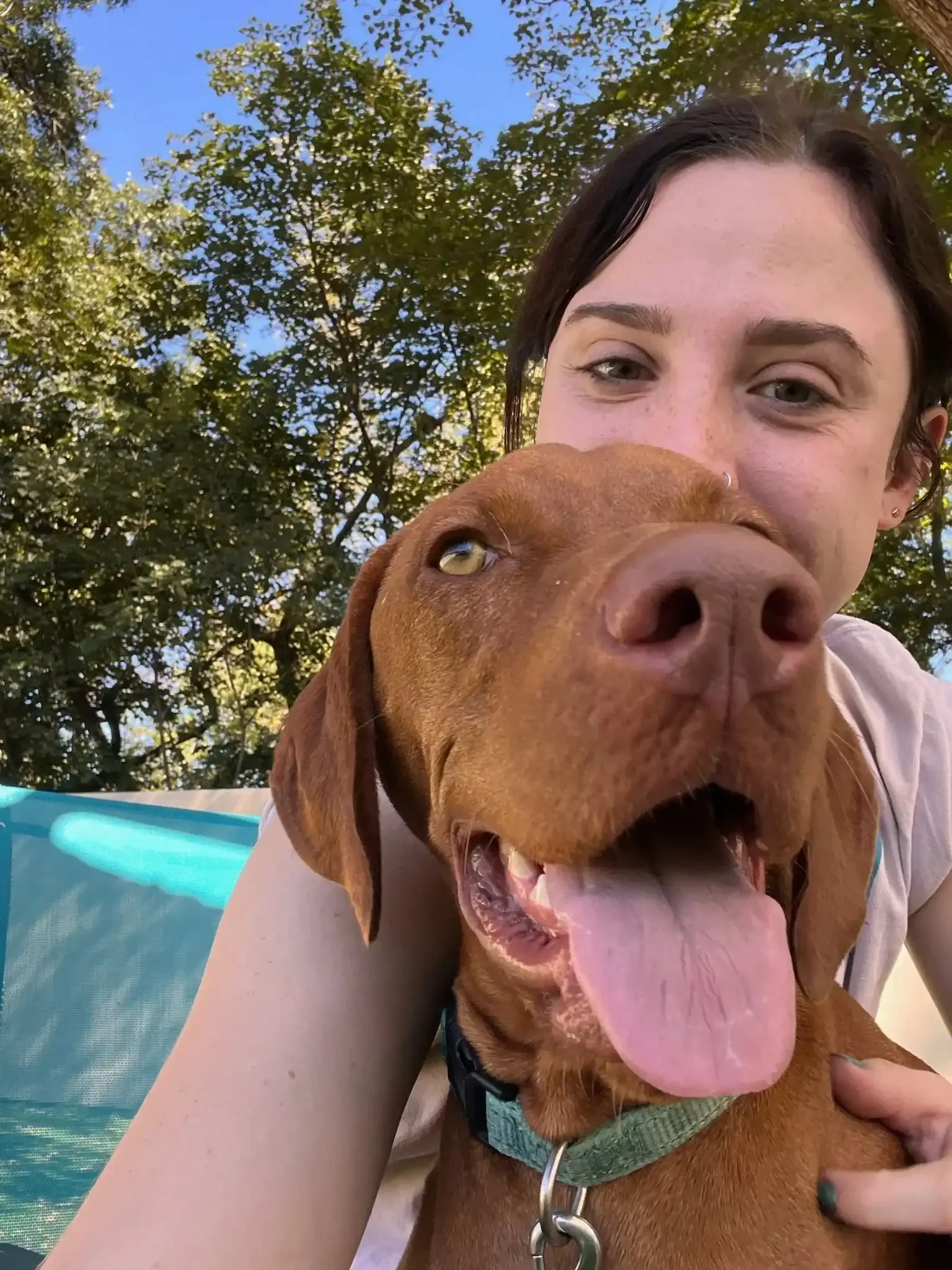 A woman with dark hair and light skin smiling while hugging a brown dog outdoors with trees and blue sky at a dog boarding location in Omaha.
