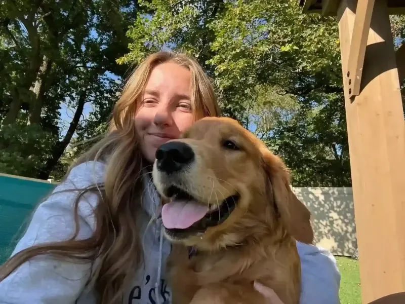A woman smiling and holding a happy golden retriever outdoors on a sunny day at a dog boarding facility in Omaha, with trees and a wooden structure in the background.
