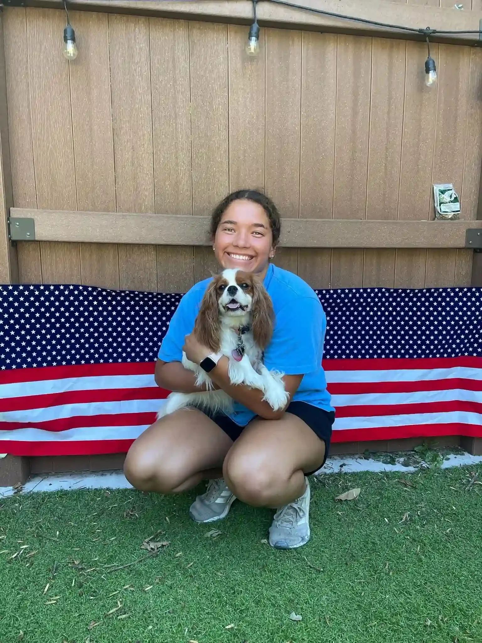 Young woman kneeling on the lawn holding a Cavalier King Charles Spaniel in a decorated outdoor play space at a dog boarding location in Omaha.