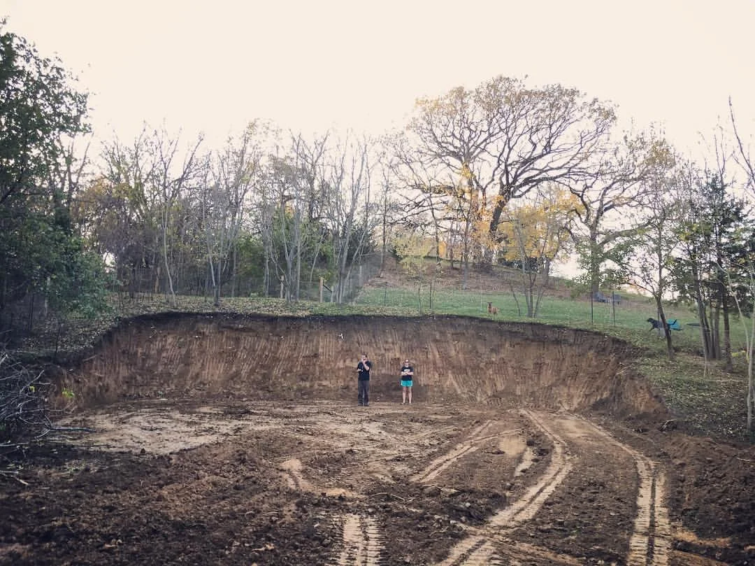 Two people standing in a large excavated hole, with a hill and trees in the background and tire tracks on the dirt in the foreground near Katie’s Kennel in Omaha, NE