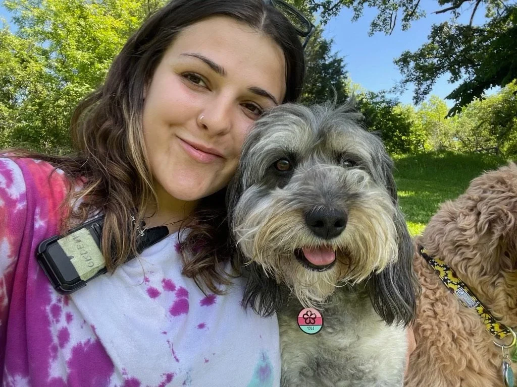 A woman with long brown hair and a nose piercing takes a selfie with two dogs outdoors on a sunny day, with trees and green grass in the background at Katie’s Kennel in Omaha, NE