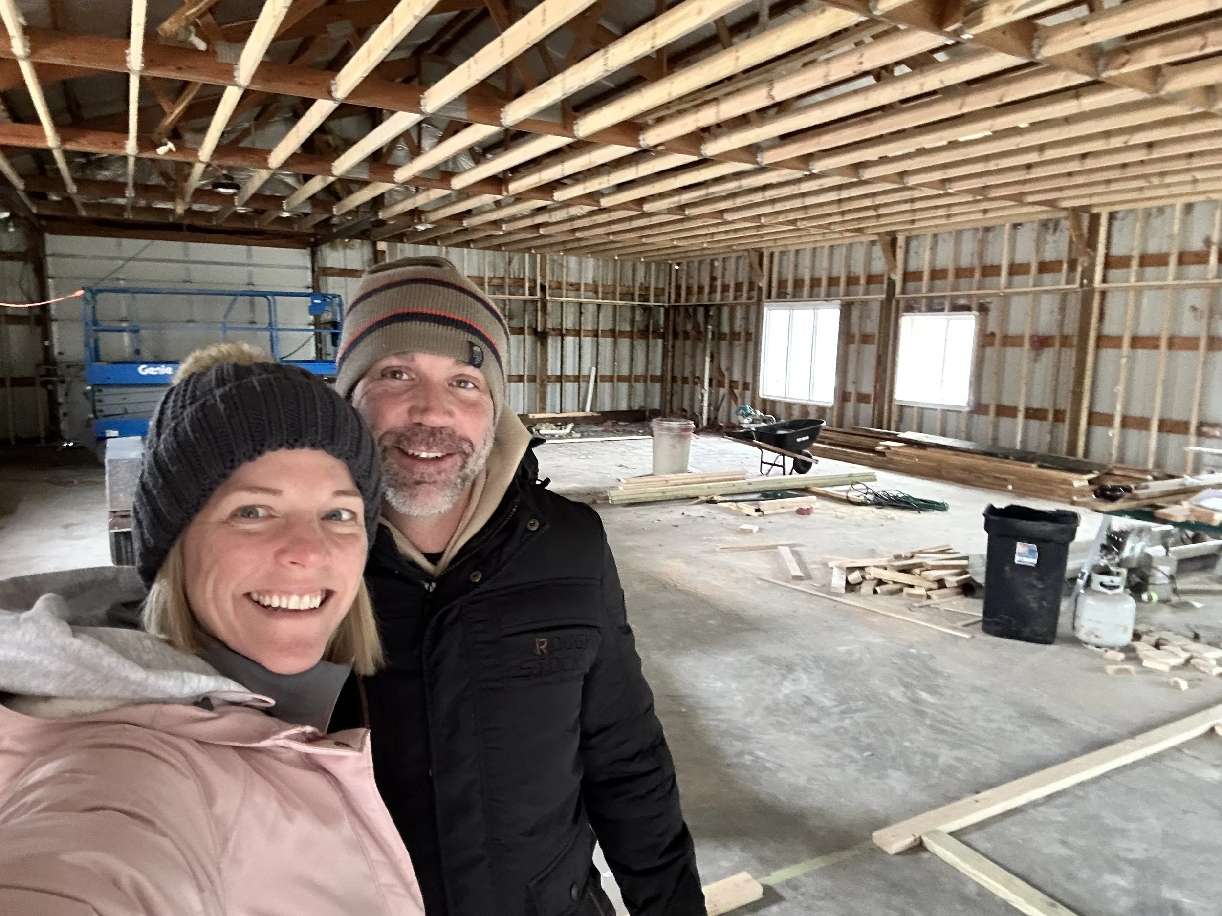 A smiling woman and man wearing winter clothing and beanies taking a selfie inside a partially constructed building with exposed wooden framing, construction materials, and tools scattered on the concrete floor near a dog boarding facility in Omaha