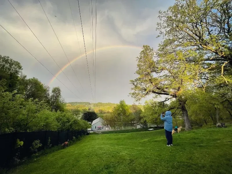 A person in a blue coat takes a picture of the cloudy sky with a rainbow shining through at a dob boarding facility in Omaha, NE.