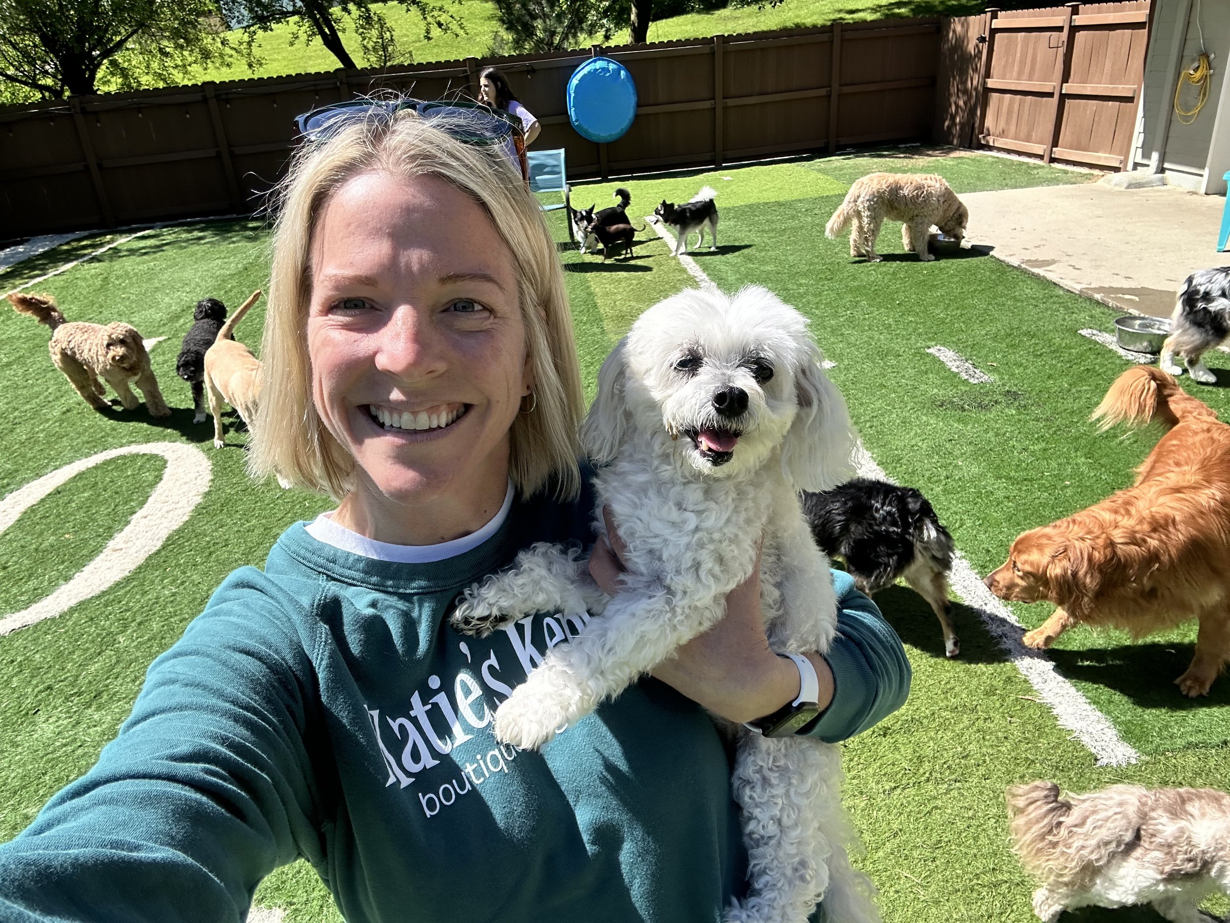 A woman smiling and holding a small white dog at a fenced outdoor dog park with multiple dogs of various breeds and colors playing on the grass at a dog boarding facility in Omaha, NE