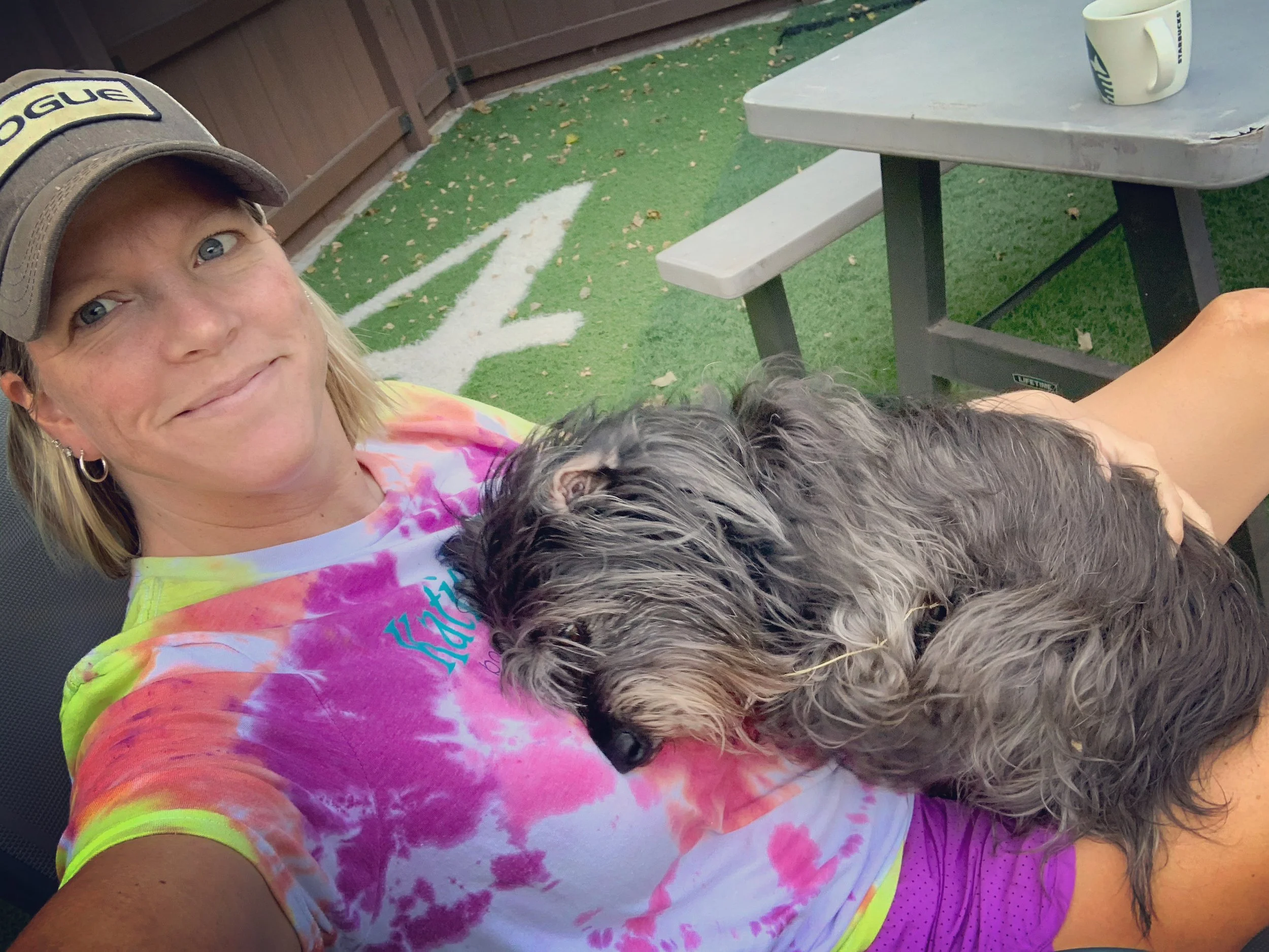 A woman wearing a tie-dye t-shirt and cap sitting outside holding a fluffy gray dog on her lap at Katie’s Kennel in Omaha, NE