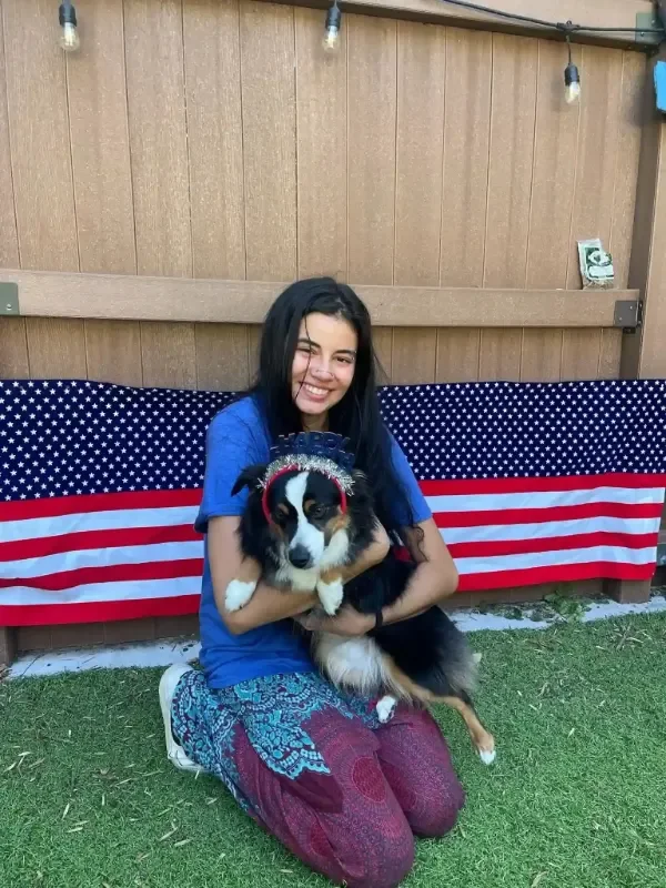 A young woman kneeling on grass while holding a black, white, and brown Australian Shepherd wearing a festive headband at an Omaha dog boarding yard decorated with lights.