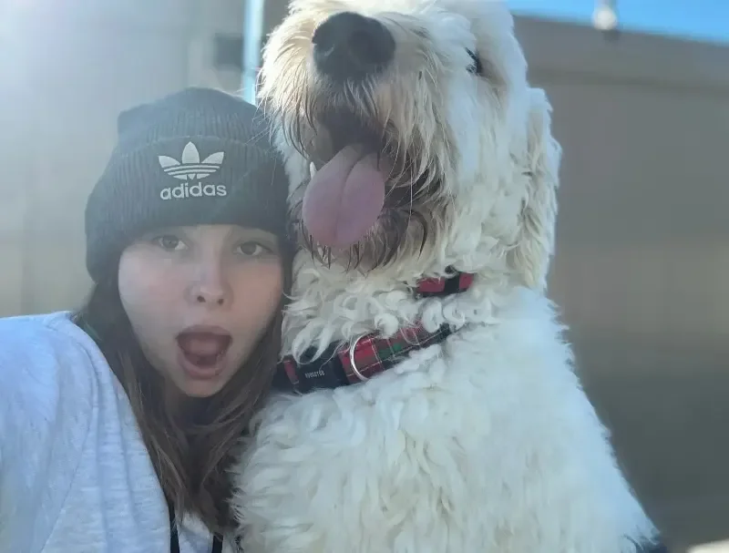 A woman in a gray hoodie and gray beanie taking a selfie with a large, fluffy white dog wearing a red plaid collar at an Omaha dog boarding center.
