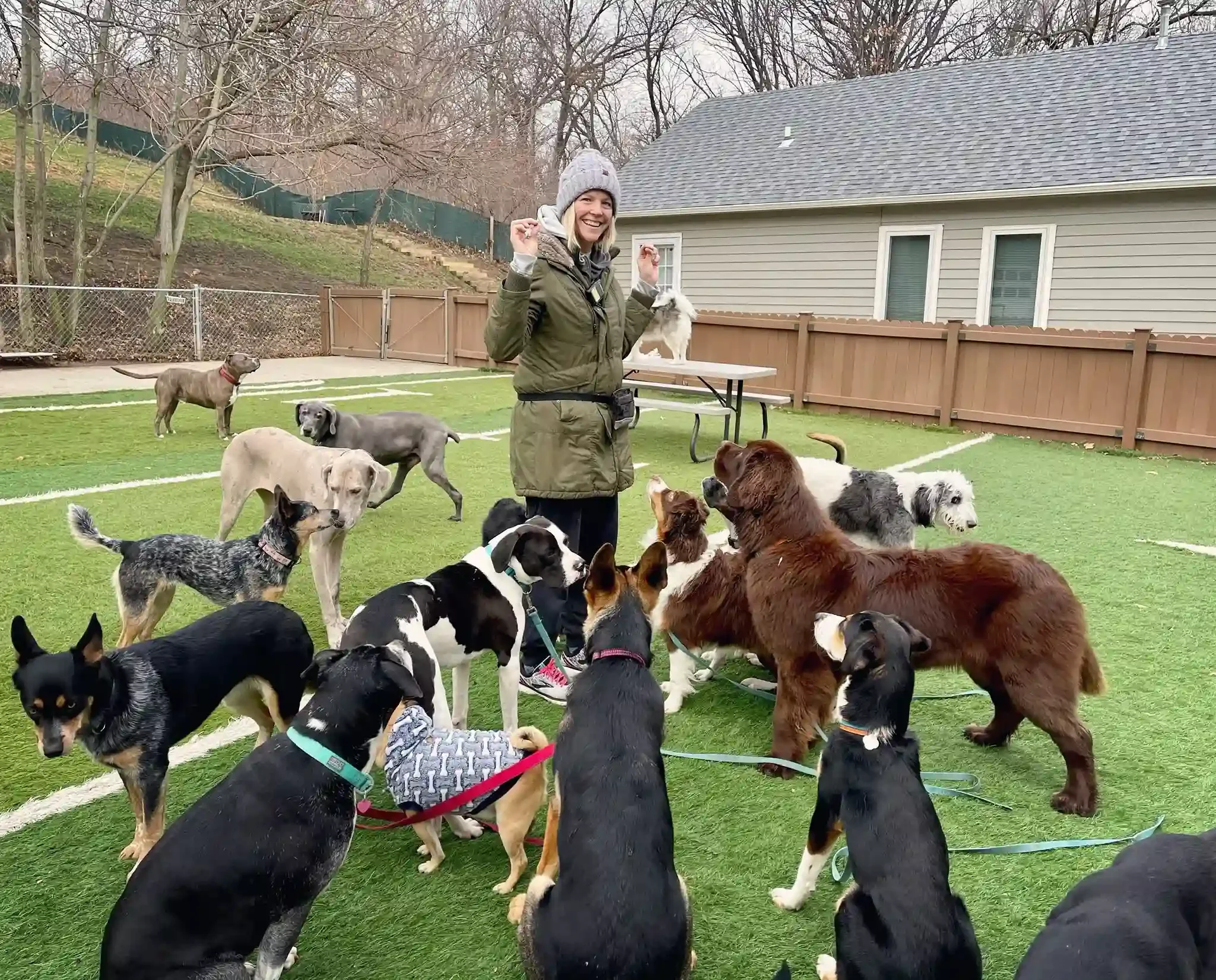 Woman surrounded by multiple dogs in a green backyard play area at an Omaha dog boarding facility.