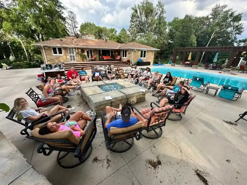 People sitting around a fire pit in a backyard with a pool, patio furniture, and a house in the background near a dog boarding facility in Omaha, NE