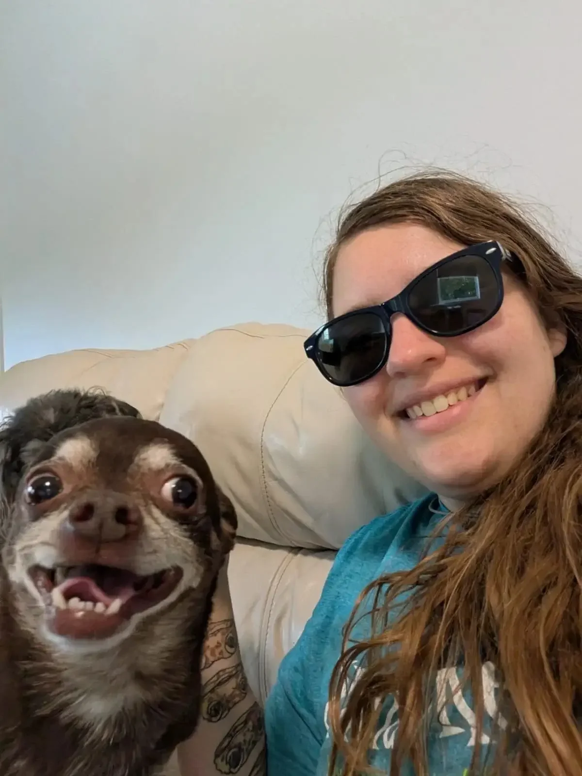 A smiling person with long hair and sunglasses taking a selfie with a happy brown and white Chihuahua relaxing on a couch inside an Omaha dog boarding lounge.