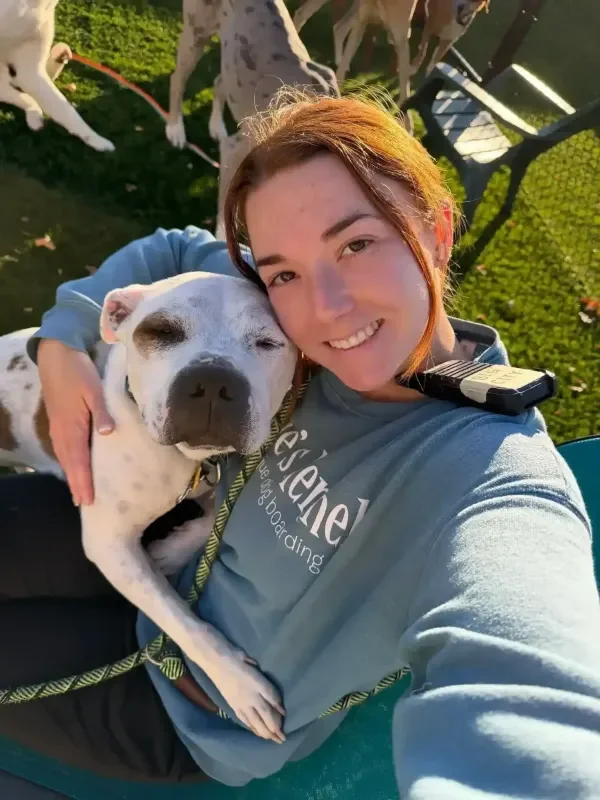 A woman taking a selfie outdoors with a happy white dog with brown spots on her lap, with other dogs playing on the grass at an Omaha dog boarding yard.