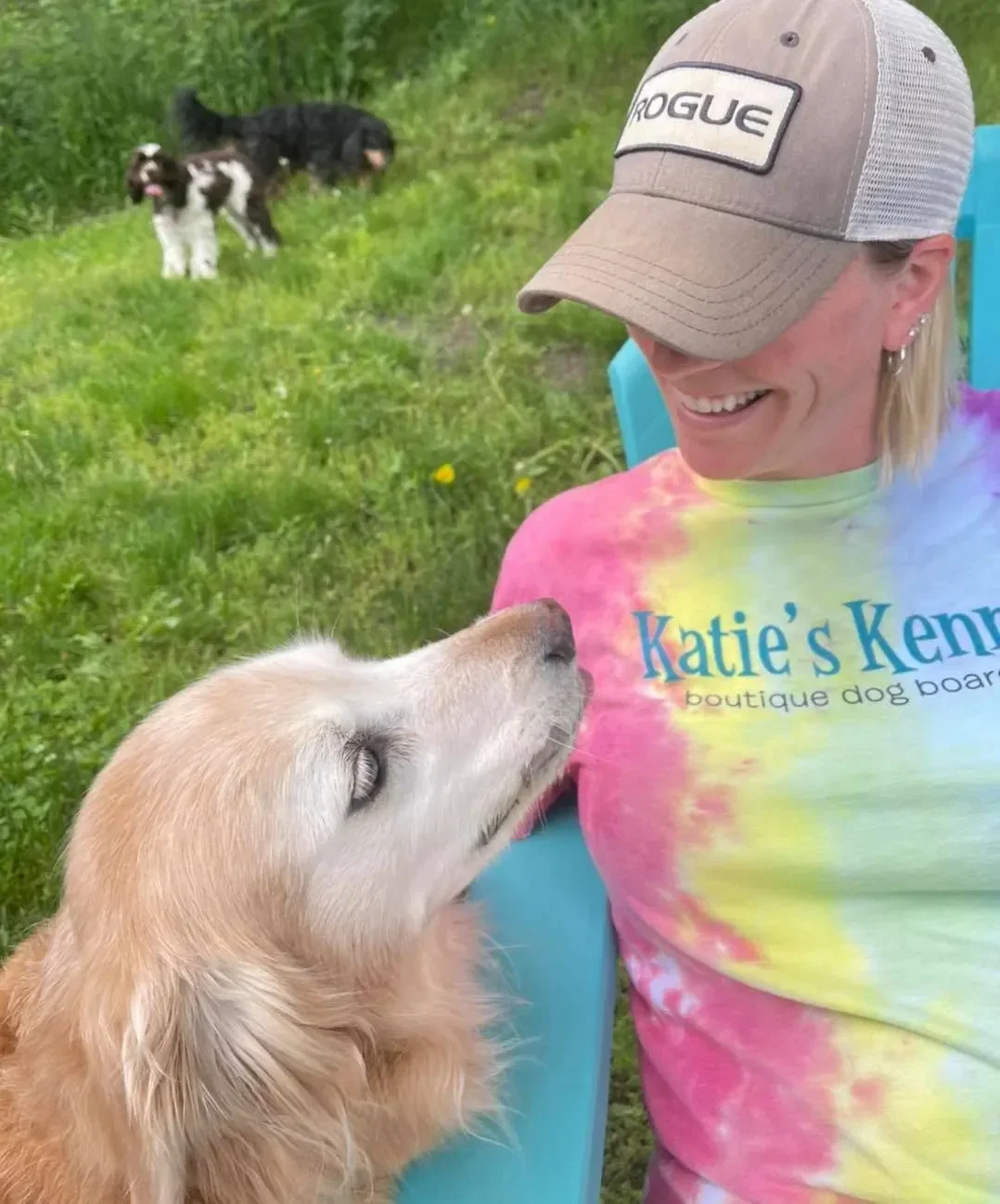 A woman in a tie-dye shirt sitting outdoors with a golden retriever puppy while other dogs relax nearby at an Omaha dog boarding yard.