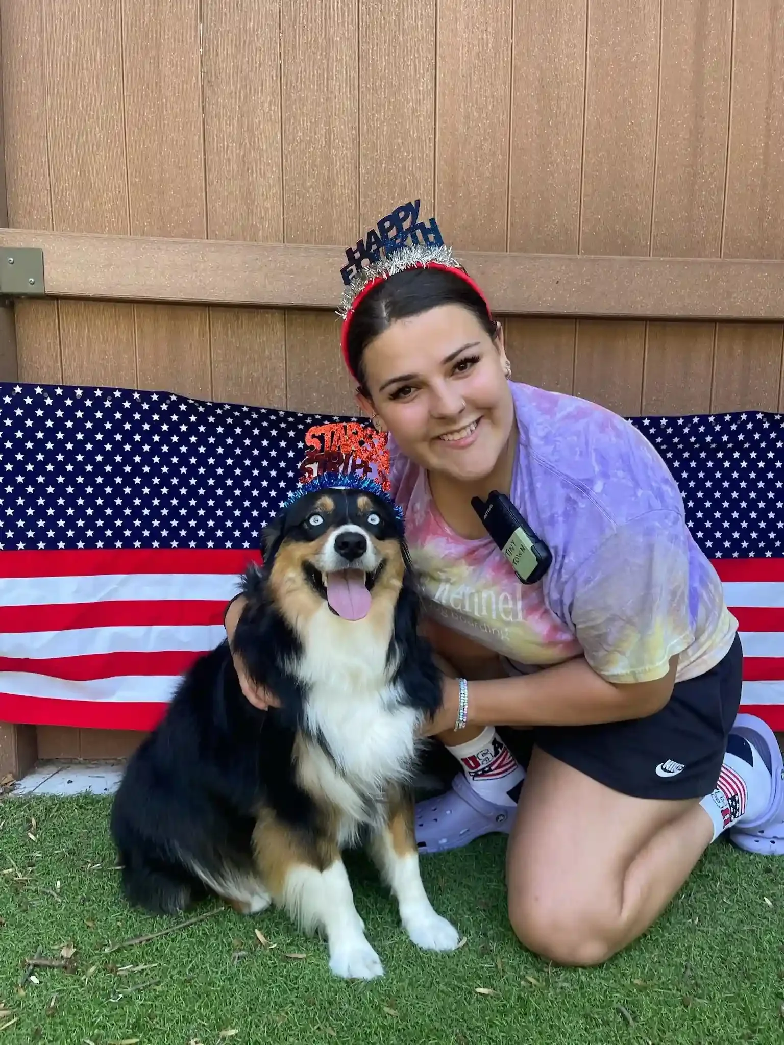 A woman with her hair up in a bun kneels next to a dark black and white dog in front of an American flag on the yard of a dog boarding center.
