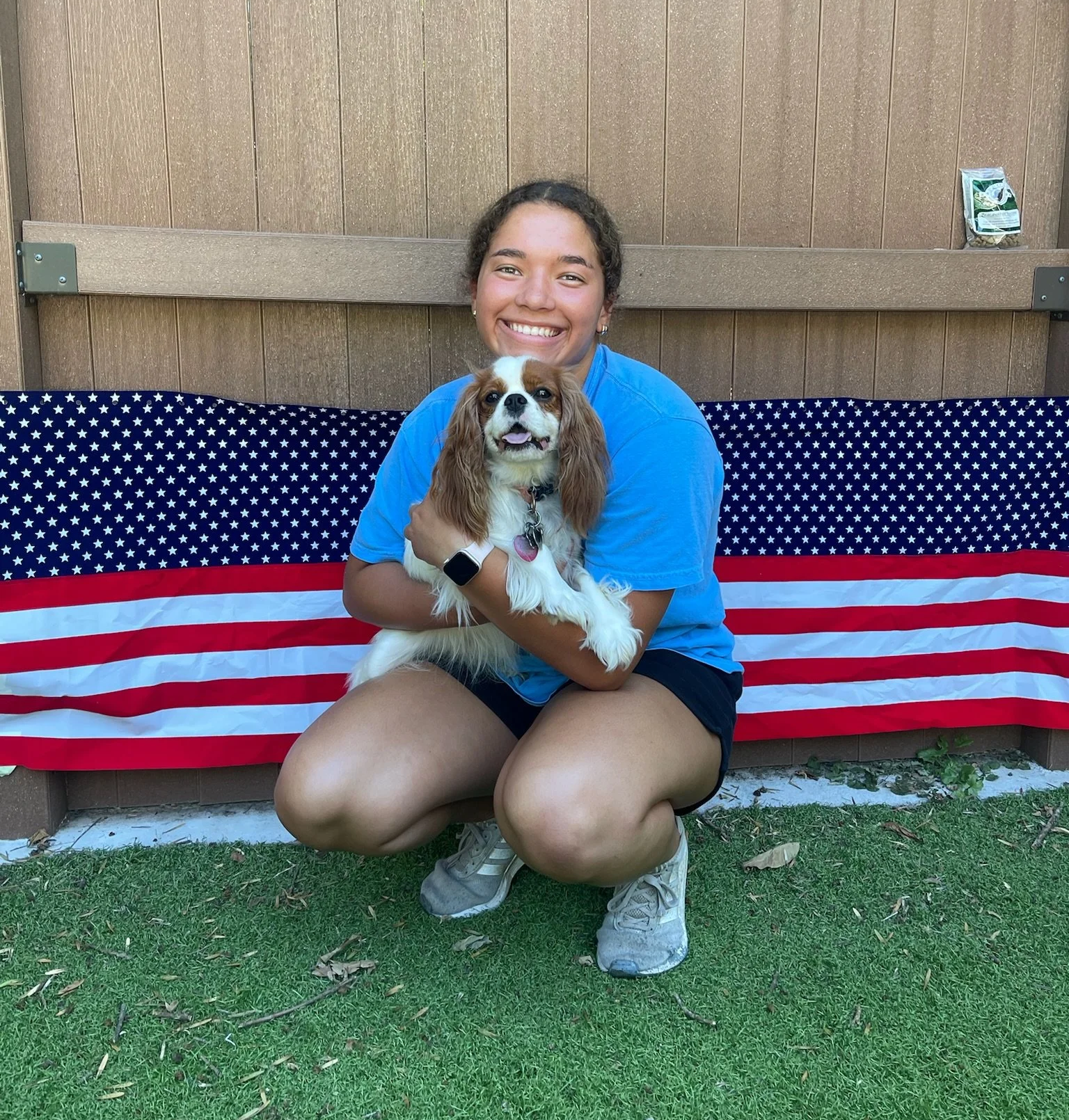 A girl sitting outdoors, holding a small dog, with a U.S. flag-themed blanket behind them and a wooden fence in the background at Katie’s Kennel in Omaha, NE