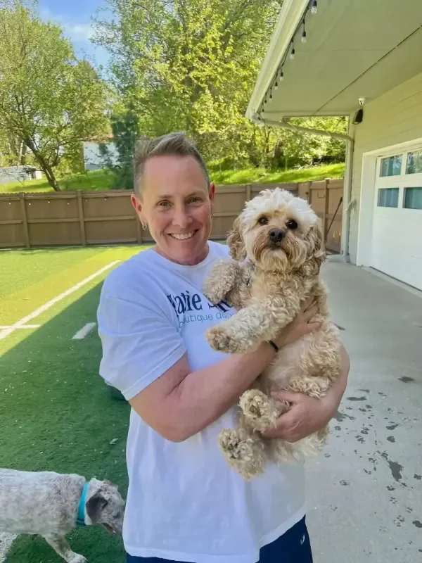 A woman smiling and holding a small, curly-haired dog outdoors in a fenced backyard play area at a dog boarding facility in Omaha, with another dog visible on the grass.