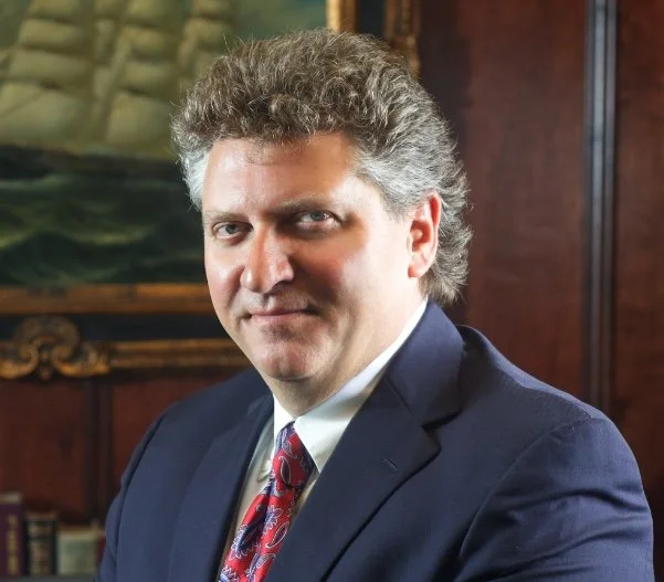 A middle-aged man with gray curly hair wearing a dark navy suit, white shirt, and patterned red tie, sitting in front of a wooden-paneled background with a painting and books.