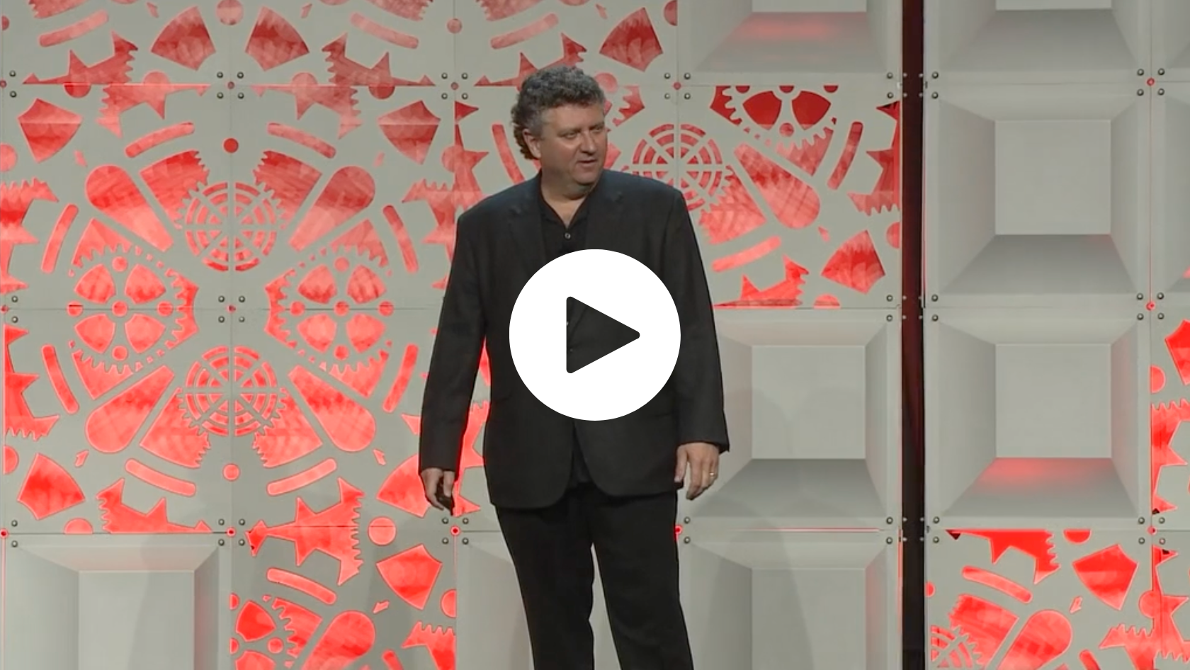A man wearing a black blazer and black pants standing on a stage with a decorative backdrop featuring geometric patterns and red lighting.