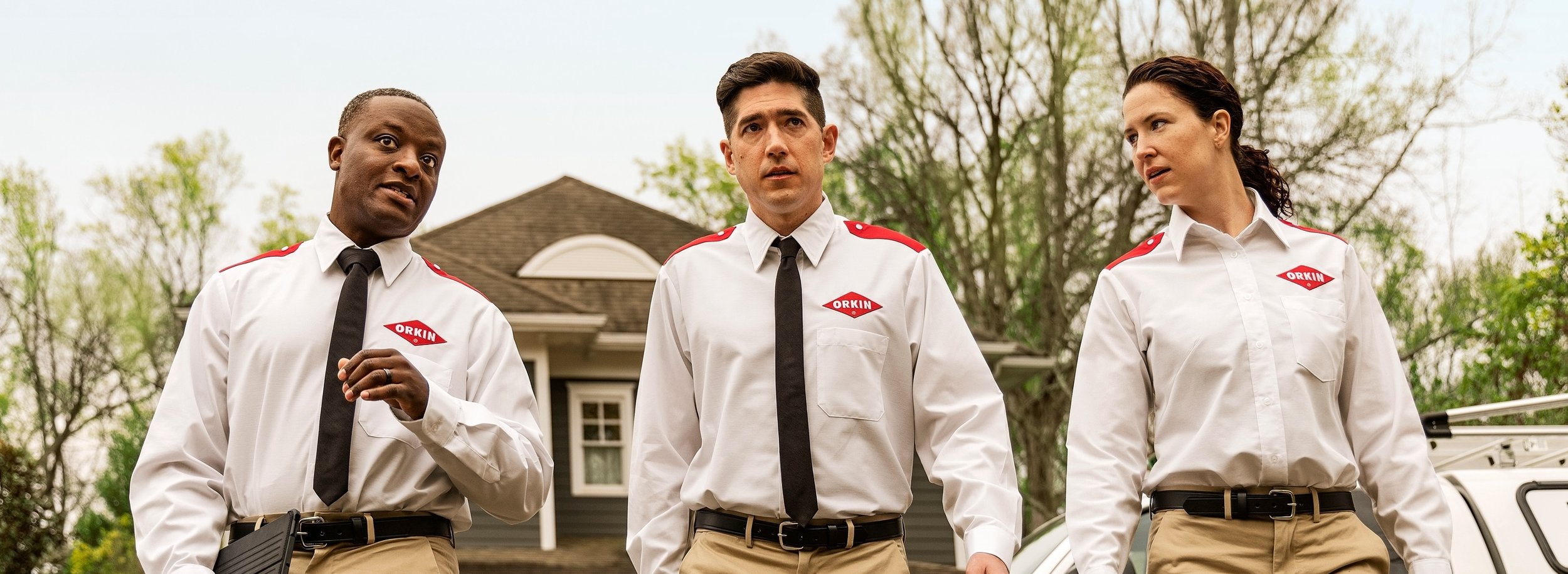 Three people dressed in white security uniforms with black ties and khaki pants standing outdoors in front of a house, with trees in the background.