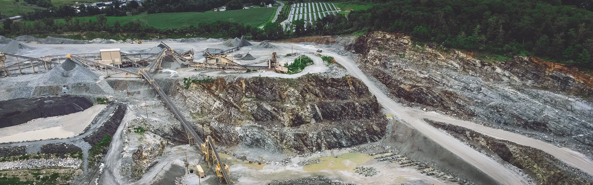 An open-pit quarry with various machinery, conveyor belts, and piles of excavated material amid a landscape with green hills and trees in the background.