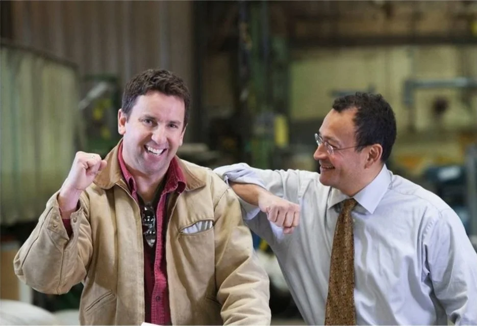 Two men are smiling and engaging in a friendly moment, with one man touching the other on the shoulder in an industrial or warehouse setting.