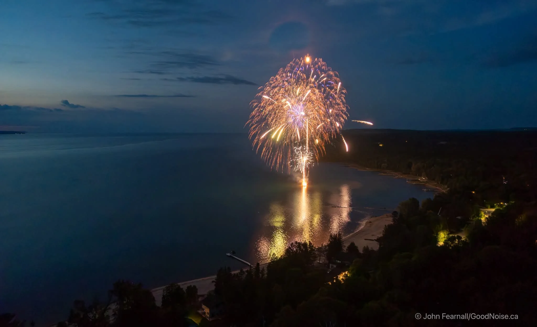 Fireworks exploding over a body of water during dusk, with trees and shoreline illuminated at night by Firedup Pyrotechnics Inc