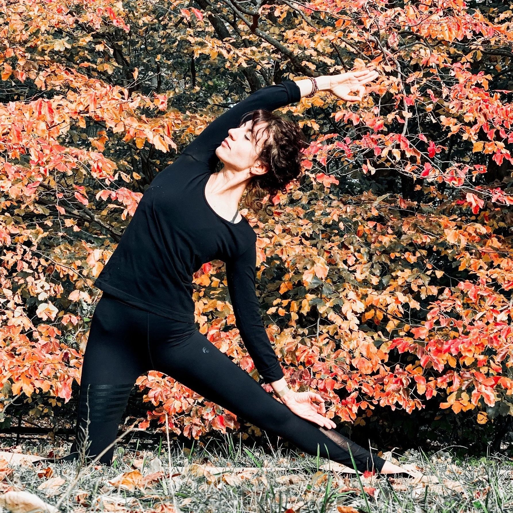 Une femme maquillée pratiquant le yoga dans un parc en automne, portant une tenue de sport noire, avec des feuilles d'automne orange et jaune en arrière-plan.