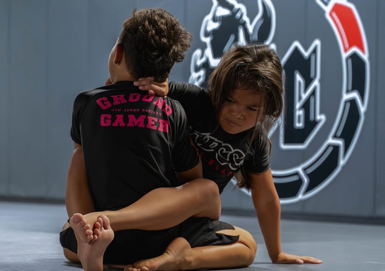 Two kids practicing Brazilian Jiu-Jitsu on a mat, one in a chokehold, in a gym with a large logo in the background.