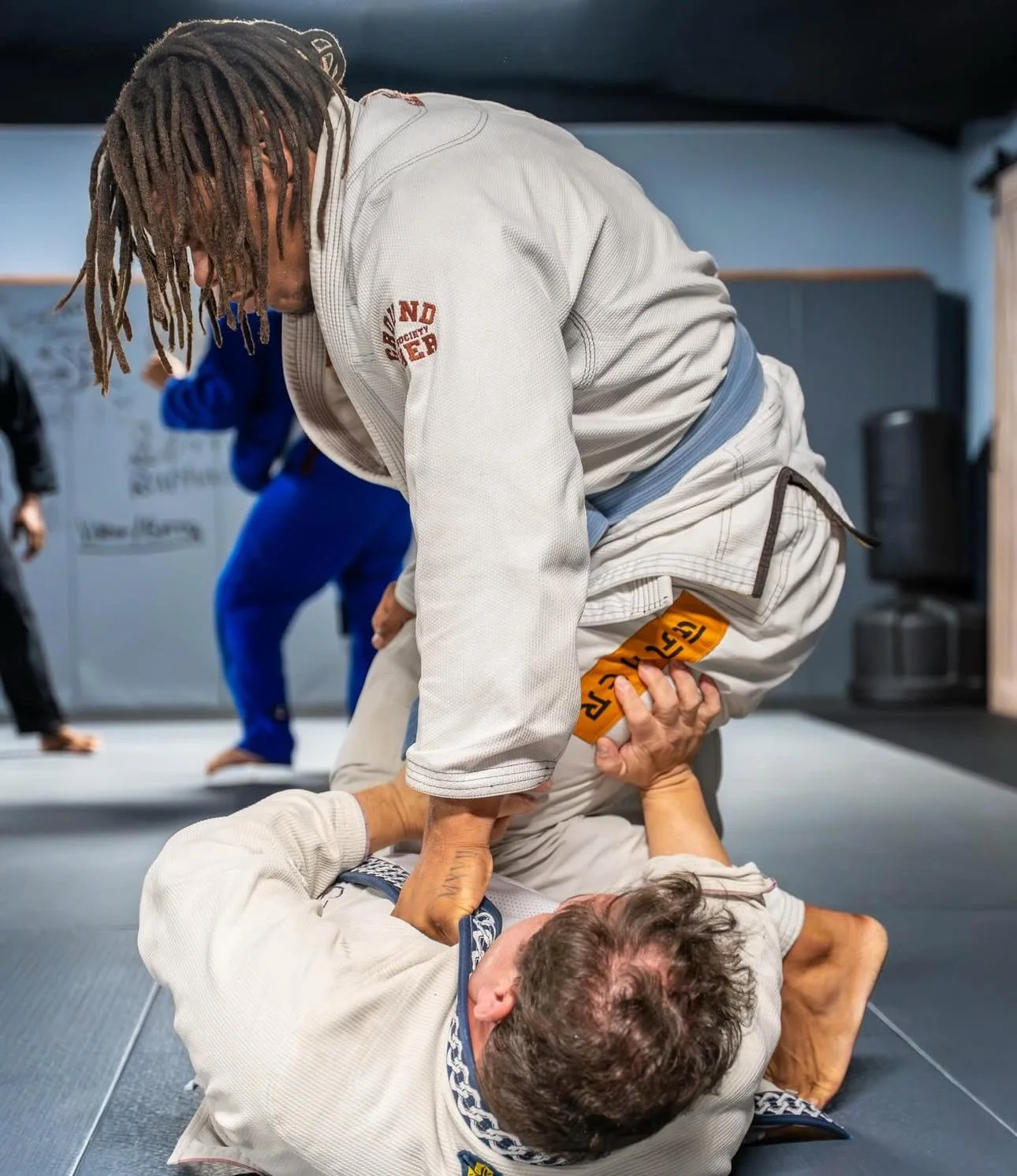 Two individuals practicing Brazilian Jiu-Jitsu in a gym; one on the ground attempting a submission hold on the other, who is in a white gi with a blue belt.