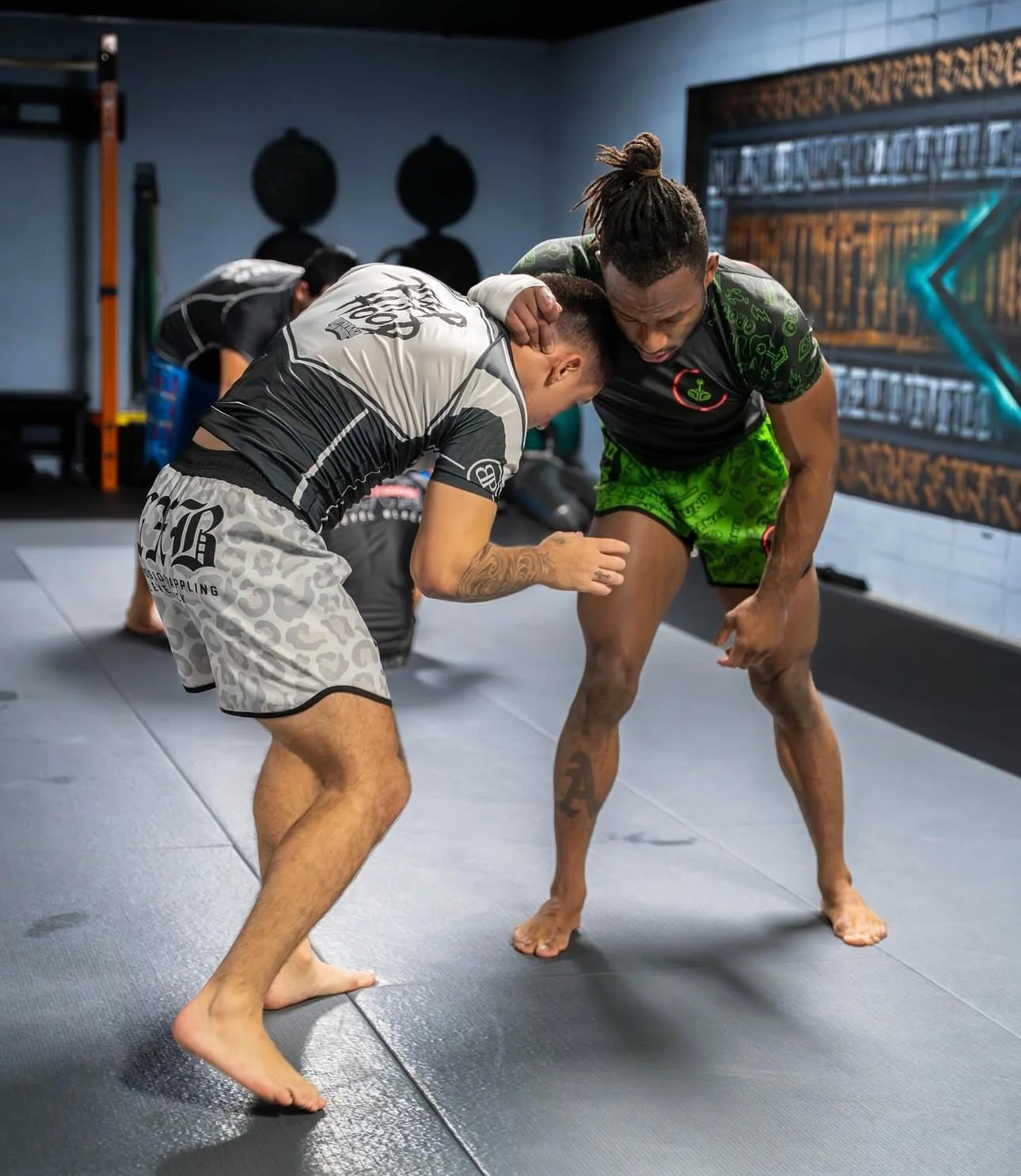 Two men practicing Brazilian Jiu-Jitsu on a mat in a gym, engaged in a grappling hold, with a person in the background.