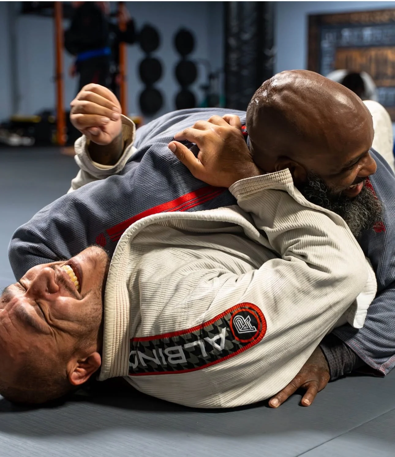 Two men engaged in a Brazilian Jiu-Jitsu match, one in a white gi and the other in a blue gi, on a gray mat inside a gym.
