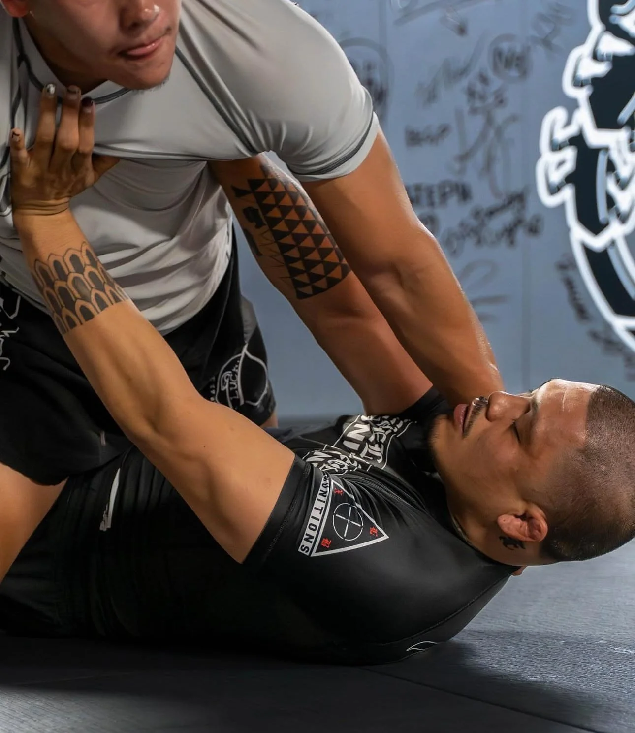 Two men practicing Brazilian Jiu-Jitsu on a mat, with one on top controlling the other, both wearing athletic clothing and tattoos visible on the person on top.