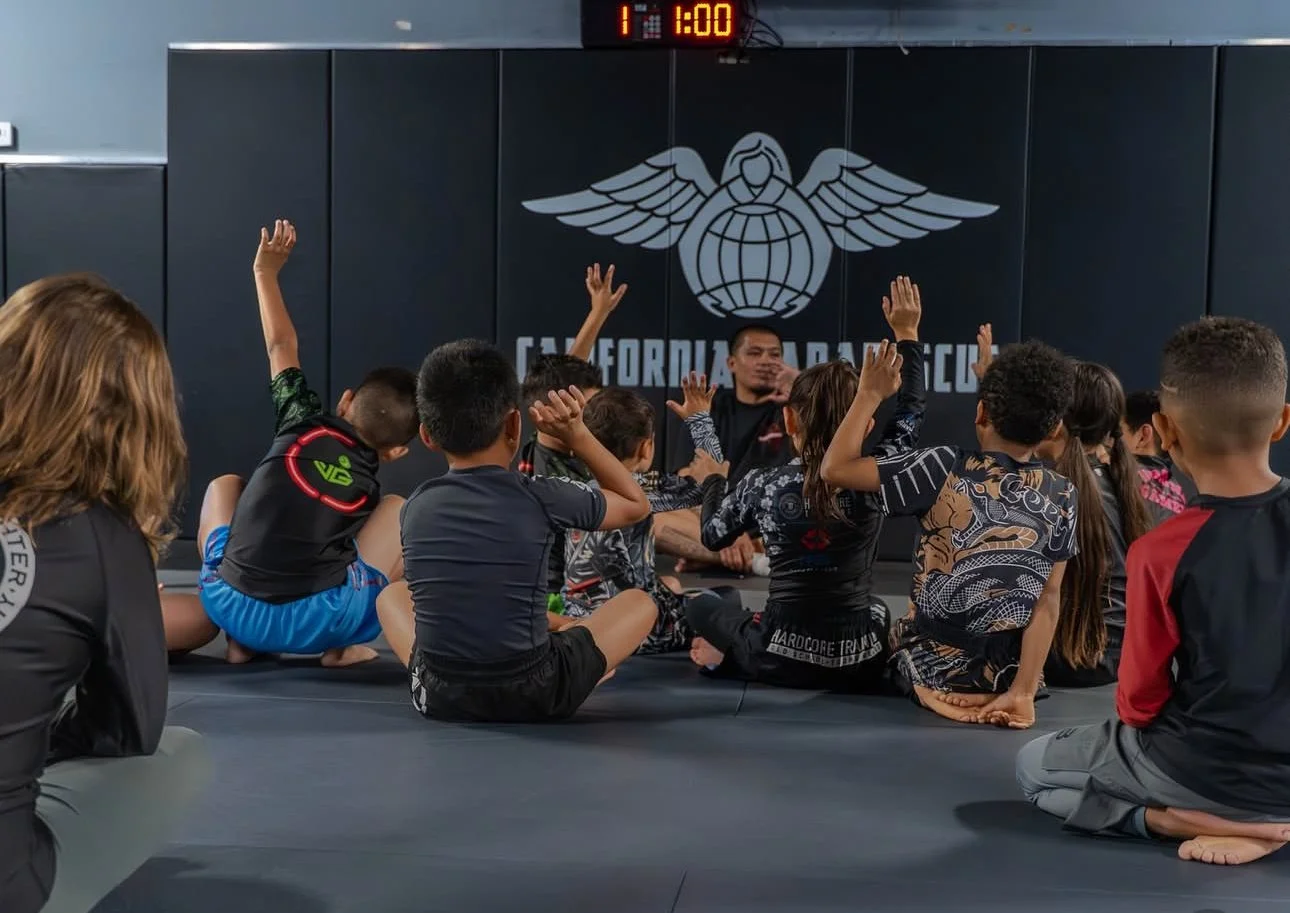 Children sitting on a martial arts gym floor, raising hands, listening to instructor, with California Martial Arts logo on black wall behind, digital clock showing 1:00 on top.