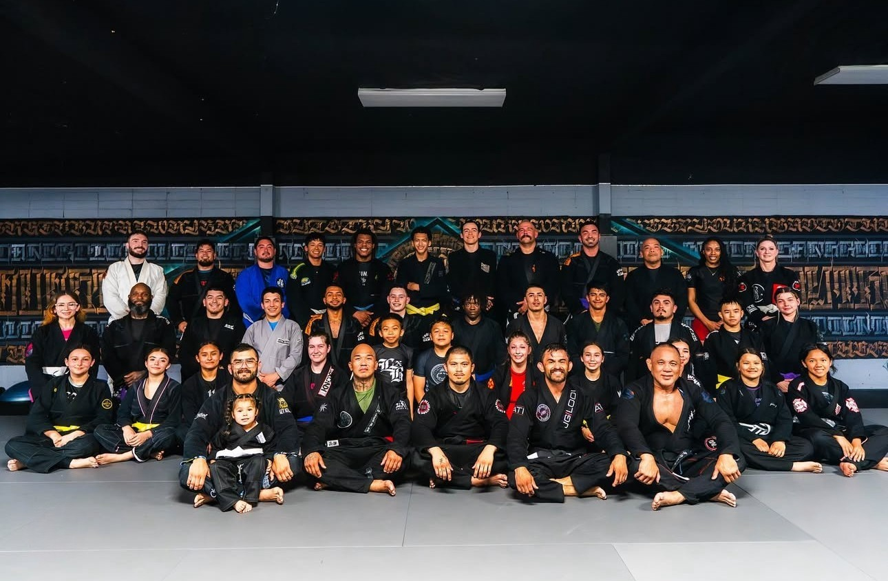 Group photo of martial artists in a dojo, wearing gis and belts, sitting and standing on a mat with a colorful mural in the background.