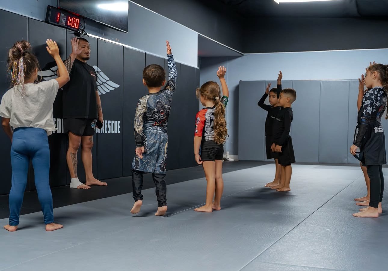 Children and instructor in martial arts class at a gym, with some children raising their hands during a demonstration or instruction.