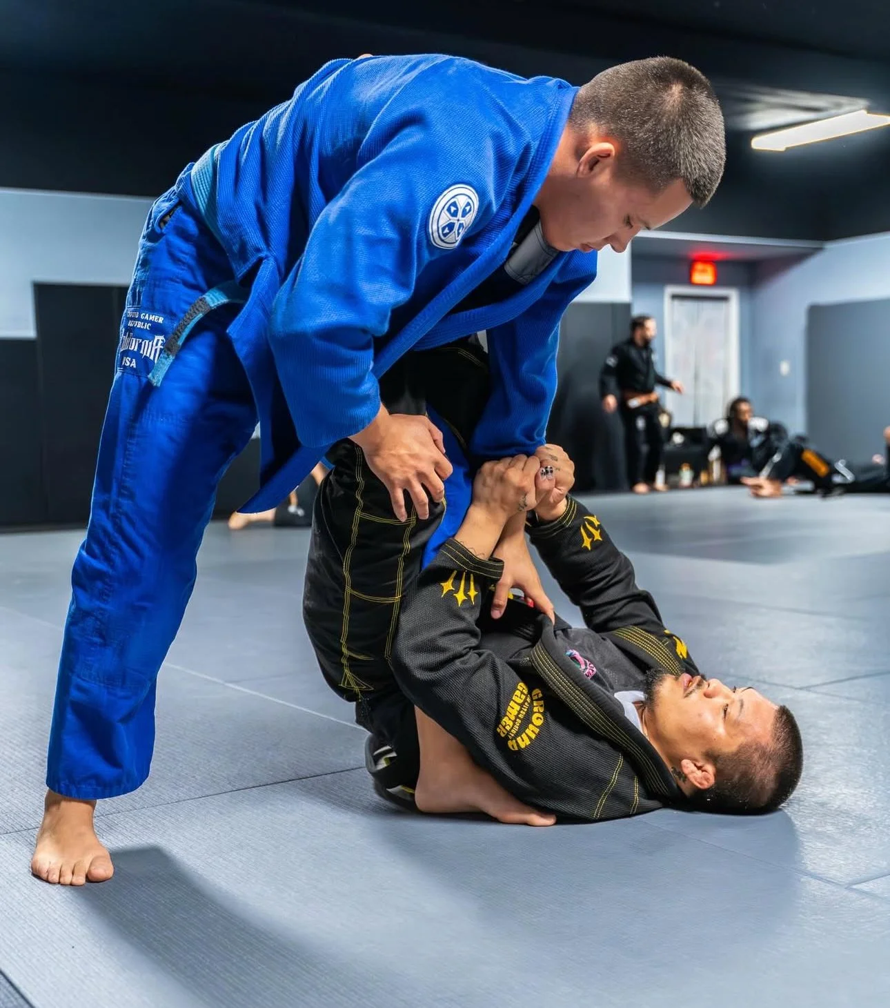 Two women practicing Brazilian Jiu-Jitsu in a gym, one in a blue gi standing over the other in a black gi on the ground, gripping her into a hold.