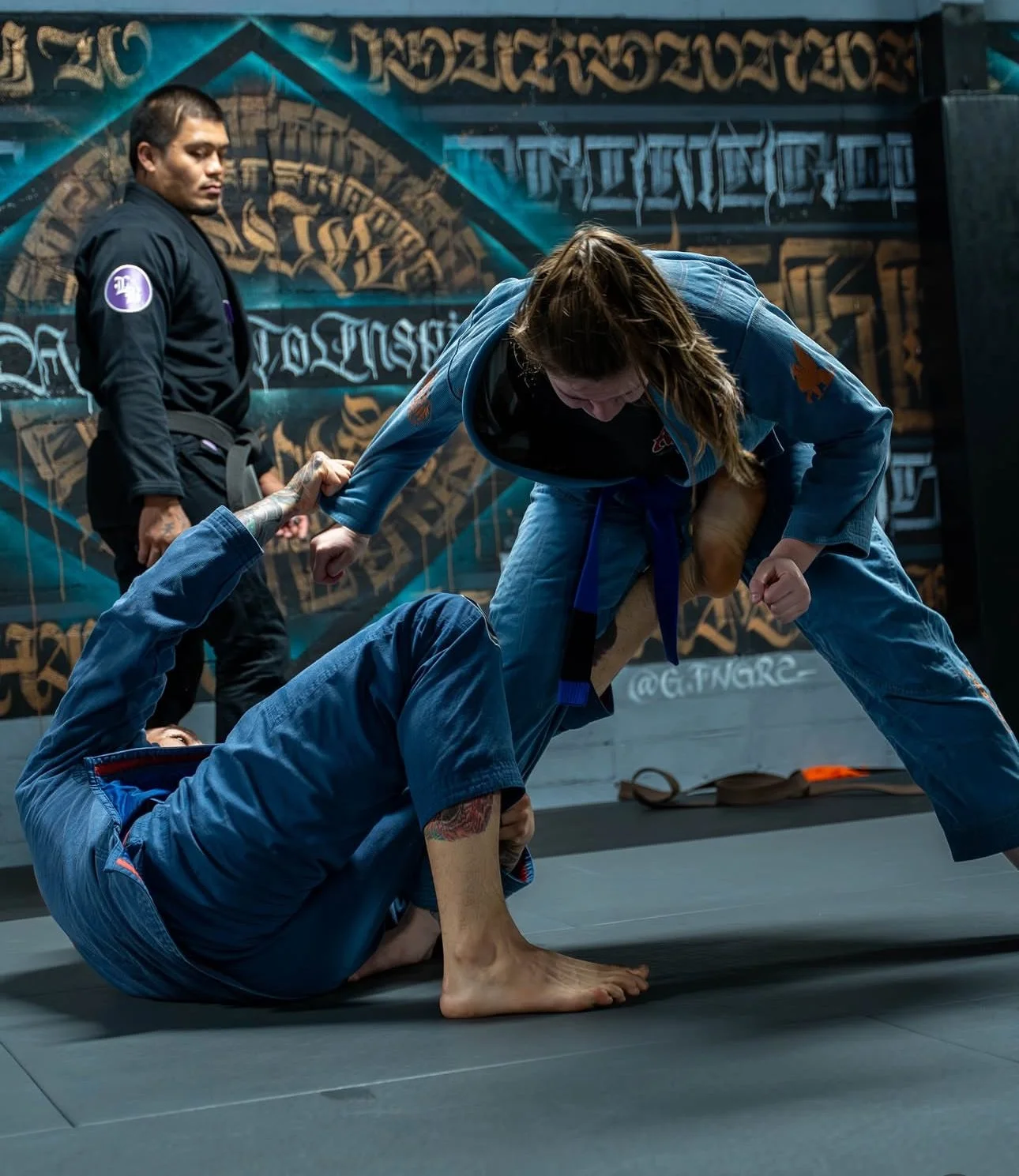 Two women practicing Brazilian Jiu-Jitsu on a mat in a gym, one woman is on the floor gripping the other woman's sleeve, while the other woman is standing and leaning over her.